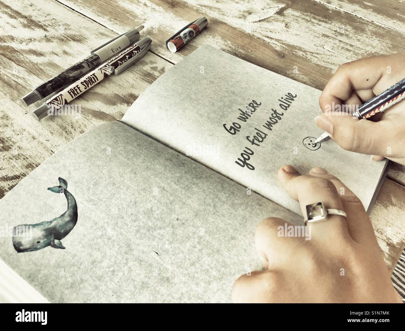 Woman drawing a smiley face in a journal open to a page with the quote Go Where You Feel Most Alive - Smartphone Captured Stock Image