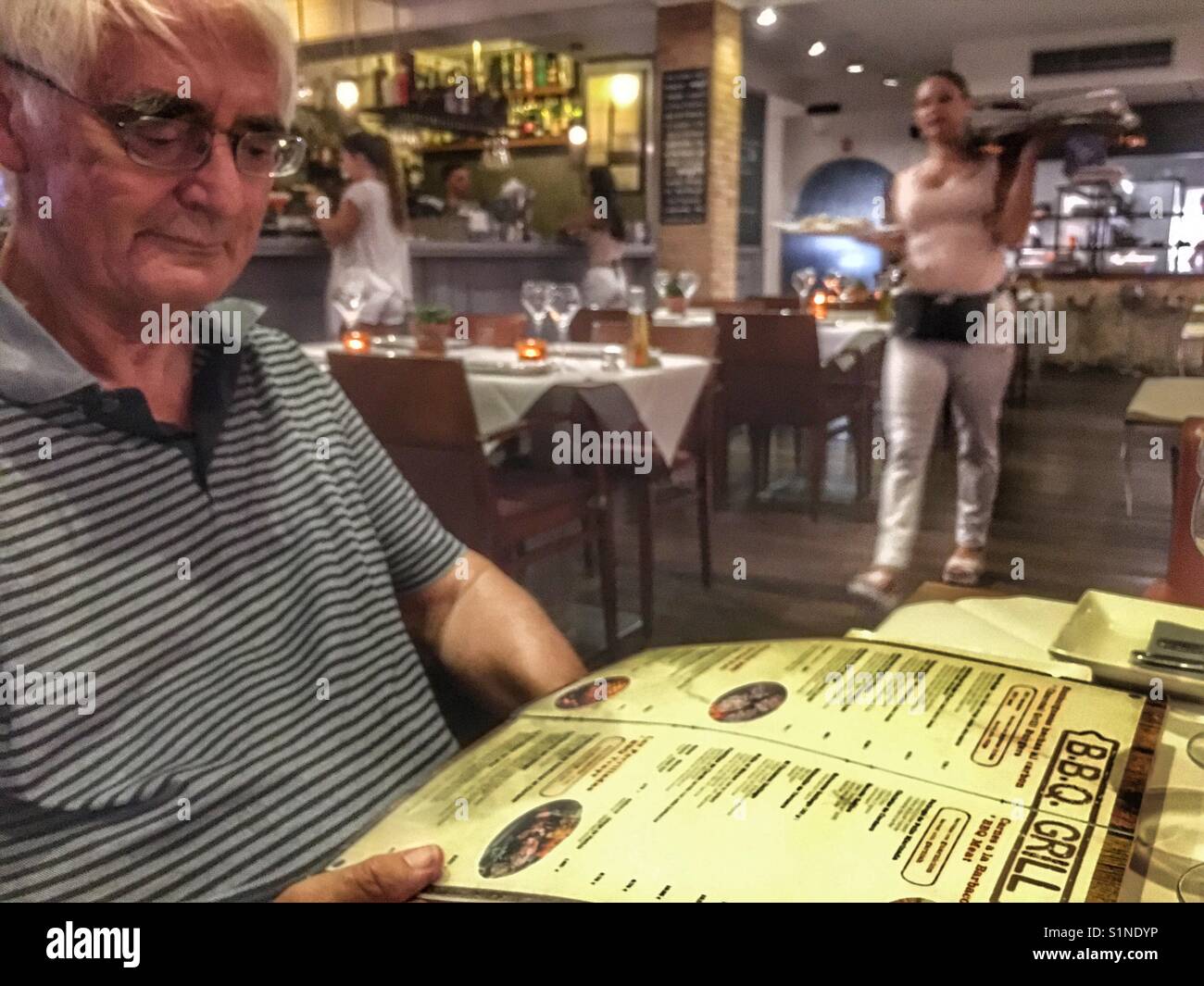 Senior man reading the menu in a restaurant with waitress approaching - Smartphone Captured Stock Image
