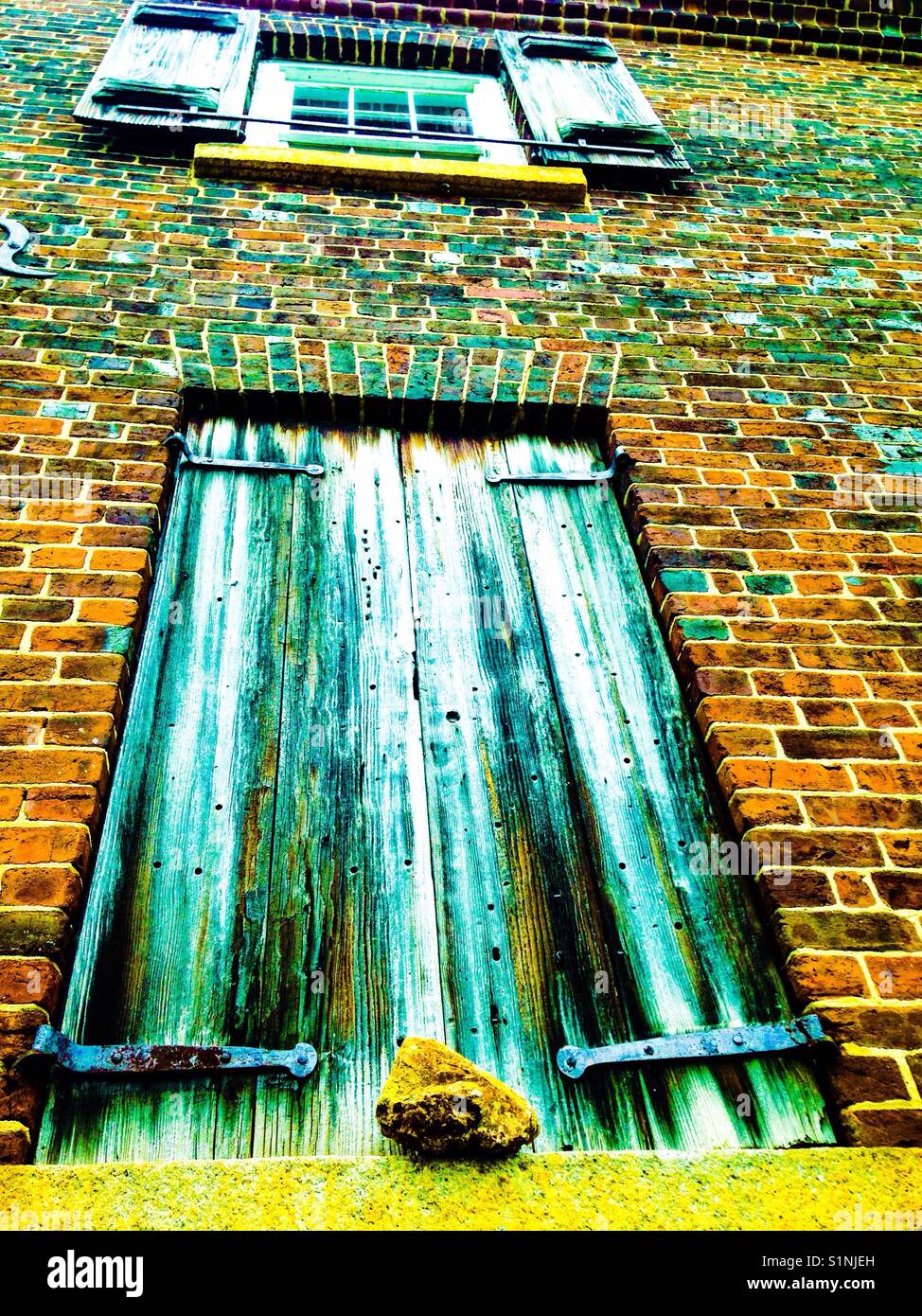 Wooden shutters on two floors in old brick building using hand forged hinges with rock holding one set shut - Smartphone Captured Stock Image