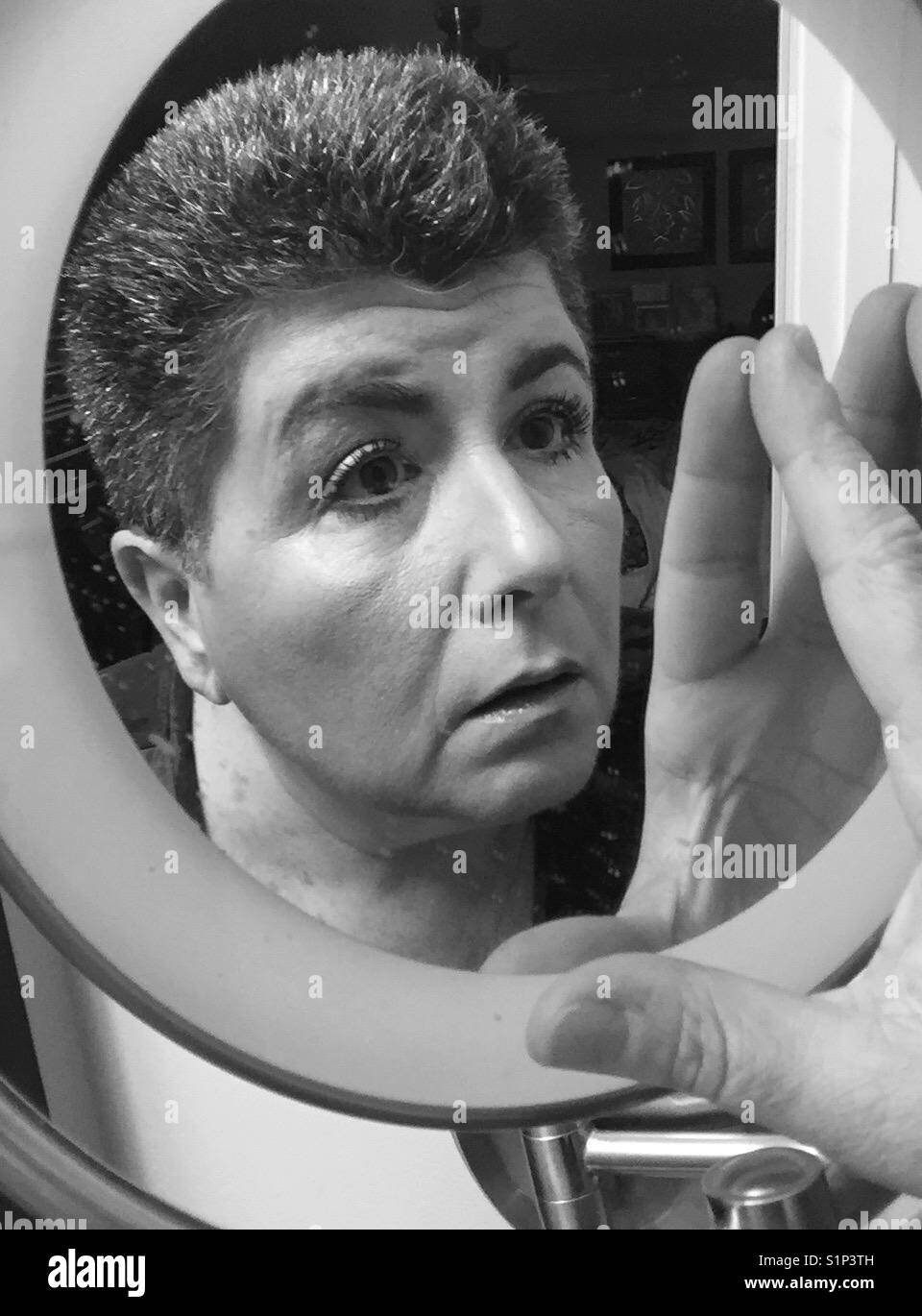 A black and white image of a dark haired middle aged woman looking into and touching a makeup mirror - Smartphone Captured Stock Image