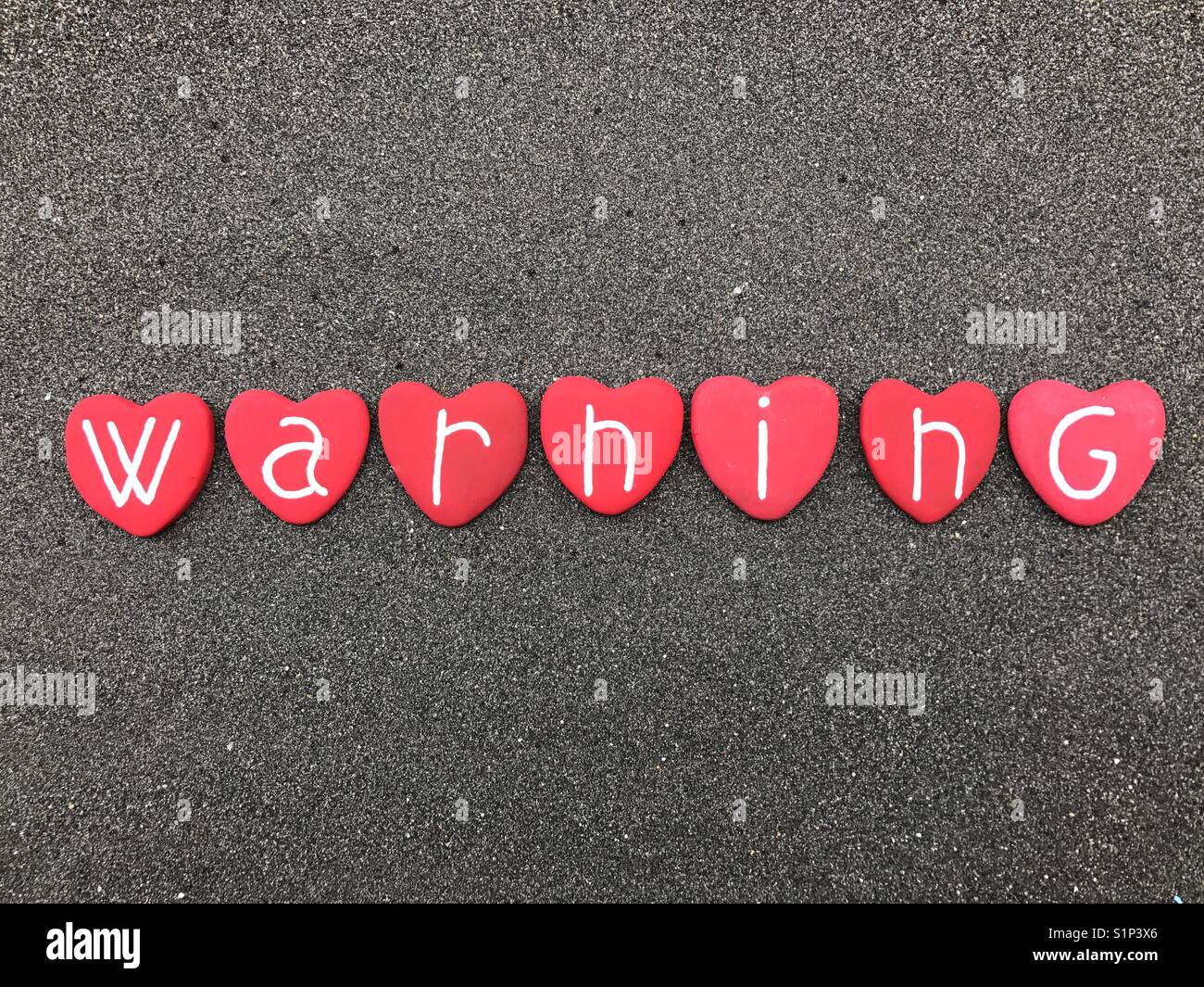 Warning text with red colored heart stones over black volcanic sand - Smartphone Captured Stock Image