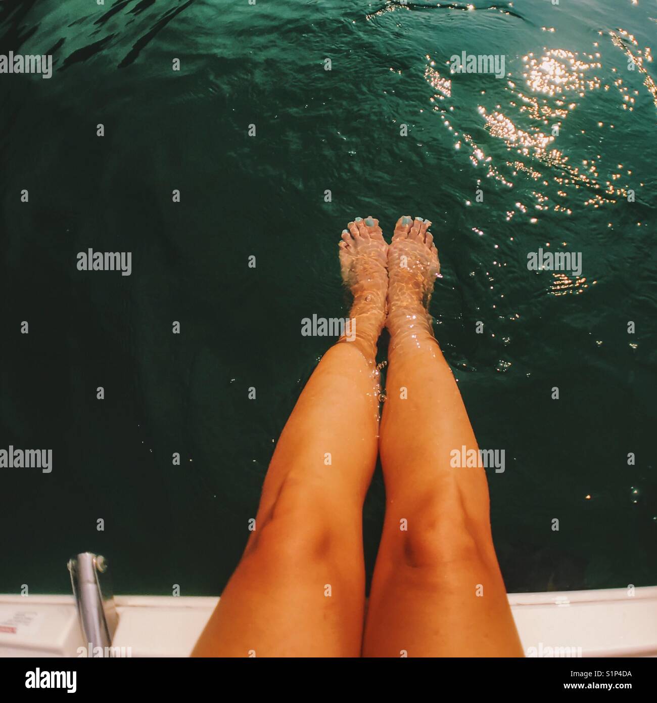 Legs of a woman sitting at the edge of a white boat, feet in the dark green water. Square crop. Stock Photo