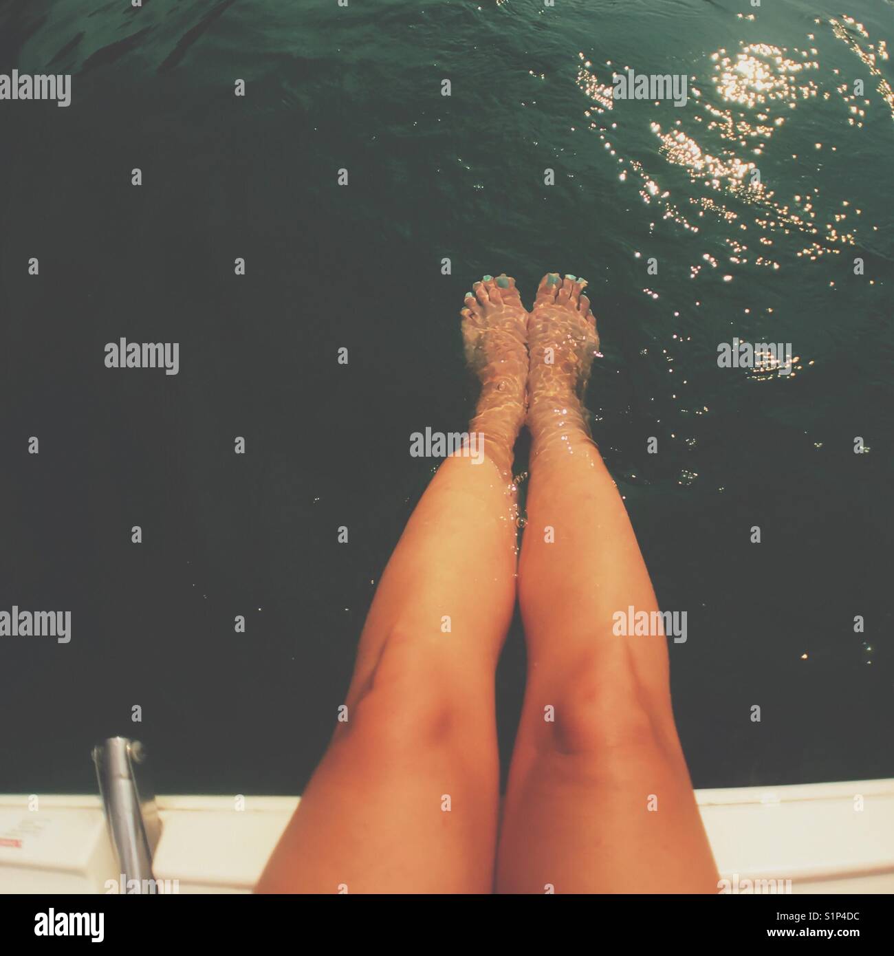 Tanned legs of woman sitting at the edge of a boat with feet submerged in water. Faded edit. Square crop. Stock Photo
