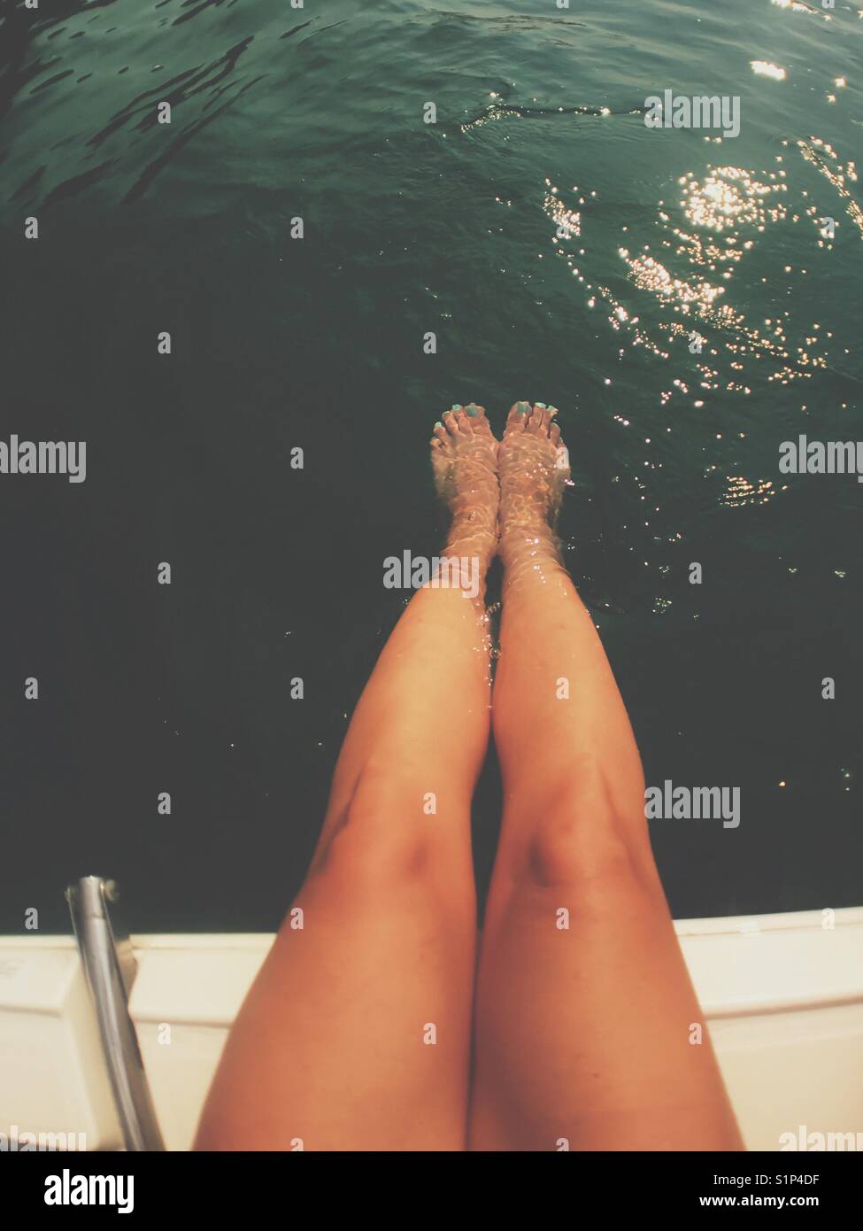 Legs of tanned woman sitting at the edge of a boat with feet submerged in water. Stock Photo