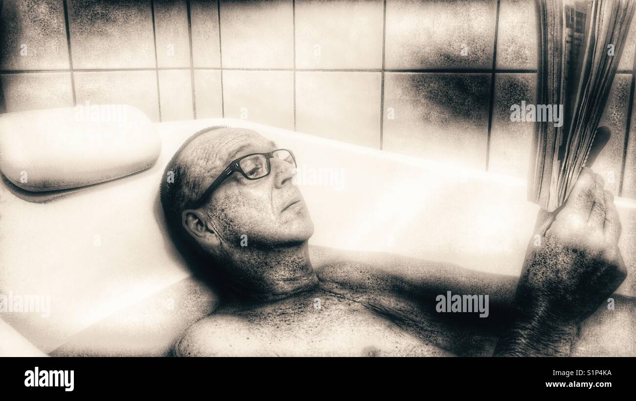 Middle aged man in glasses reclining reading a book in the bath - Smartphone Captured Stock Image