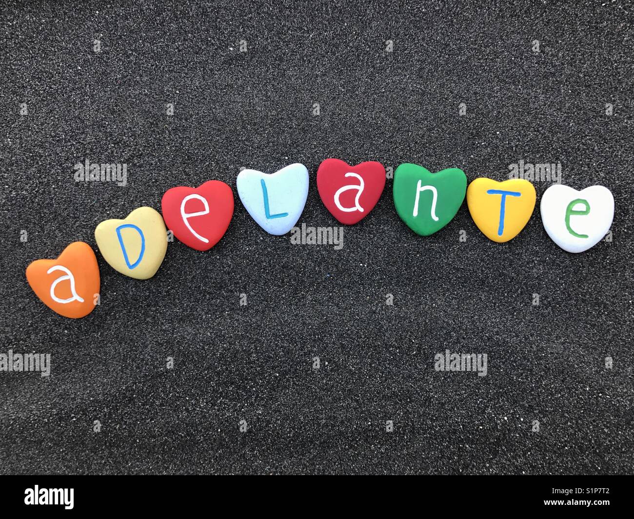Adelante, spanish word meaning ahead composed on black volcanic sand with colored stone hearts Stock Photo