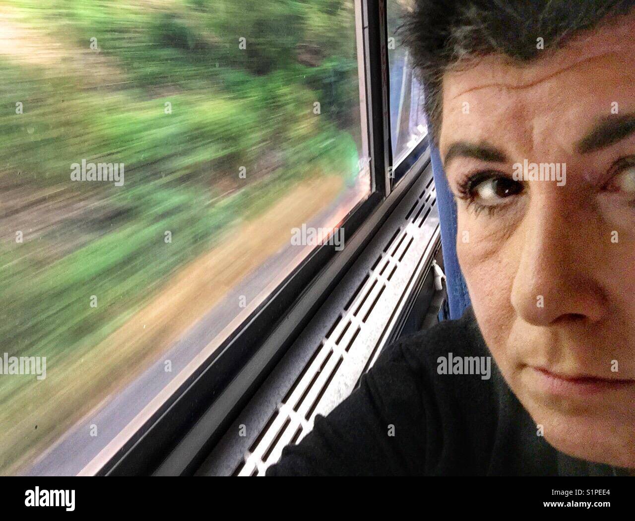 A portrait of a middle aged woman with dark hair who is riding on a train.  The outdoors is seen out the window in motion. - Smartphone Captured Stock Image