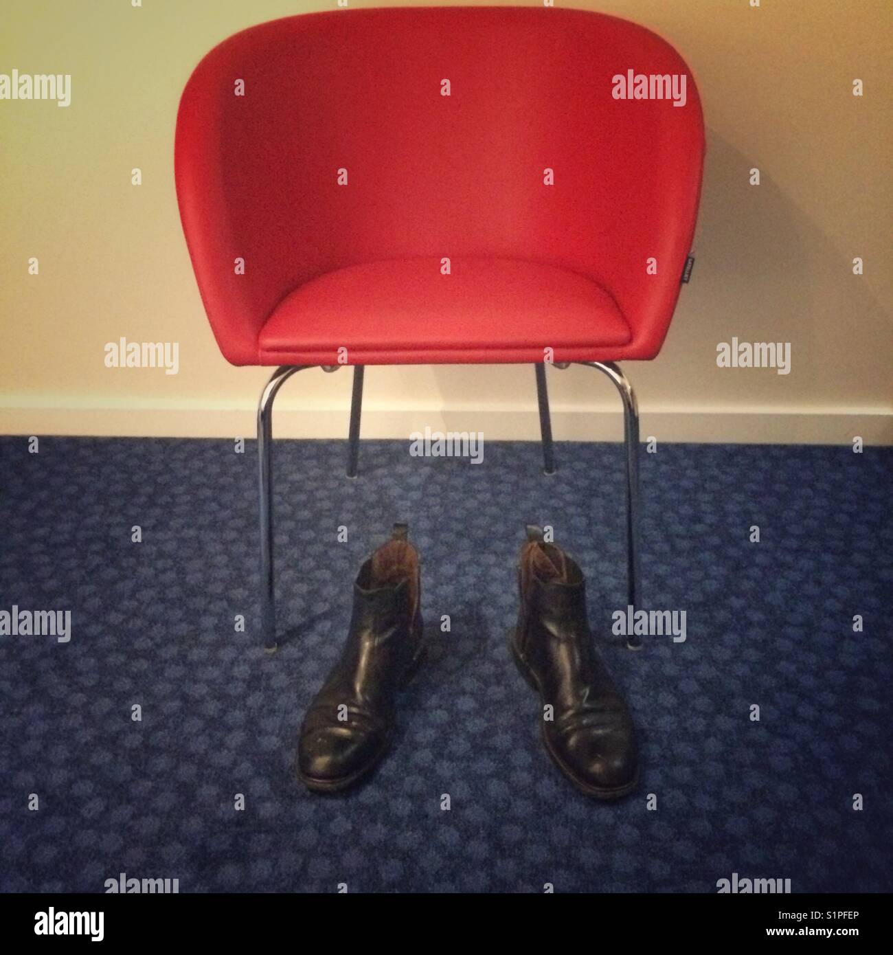 Pair of worn black Chelsea type men’s fashion boots in front of a red vinyl covered chair. Stock Photo