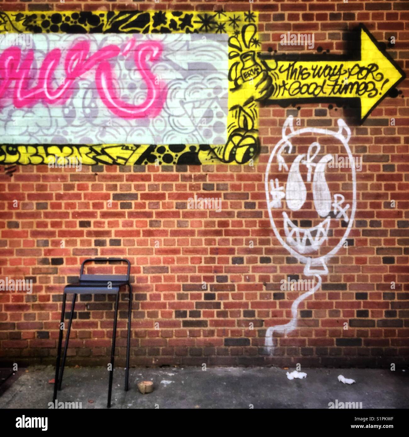 Colorful graffiti on a red brick wall that says “this way to good times” and that has a comical face painted below it.  A tall chair sits against the wAll - Smartphone Captured Stock Image