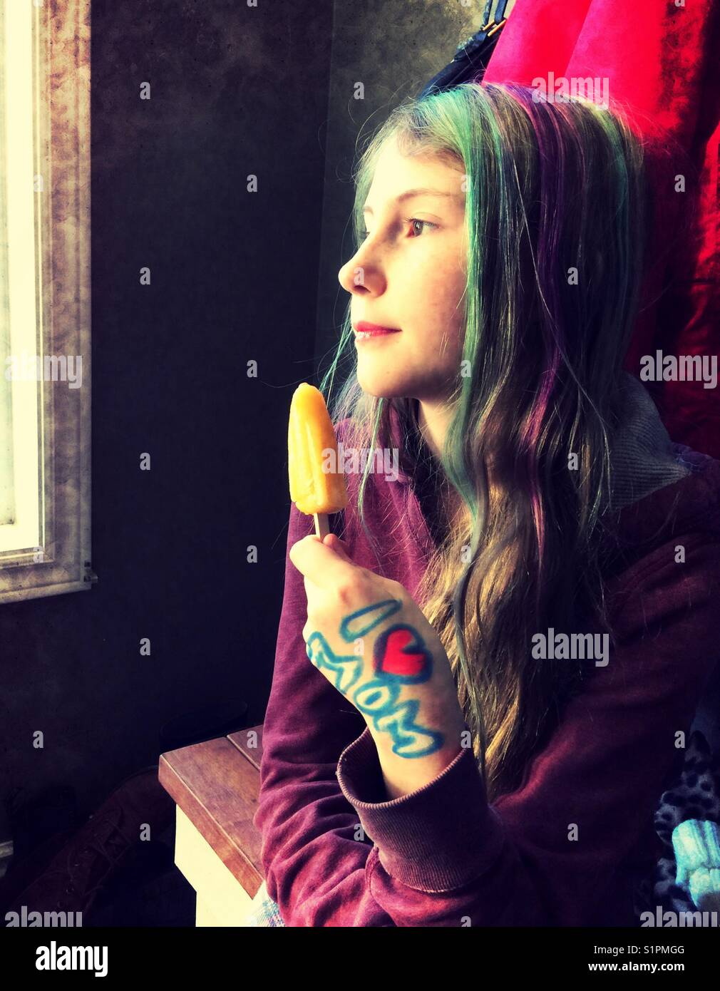 Preteen girl with dyed hair and 'I ❤️MOM' drawn on hand daydreaming by window while holding orange frozen dessert Stock Photo