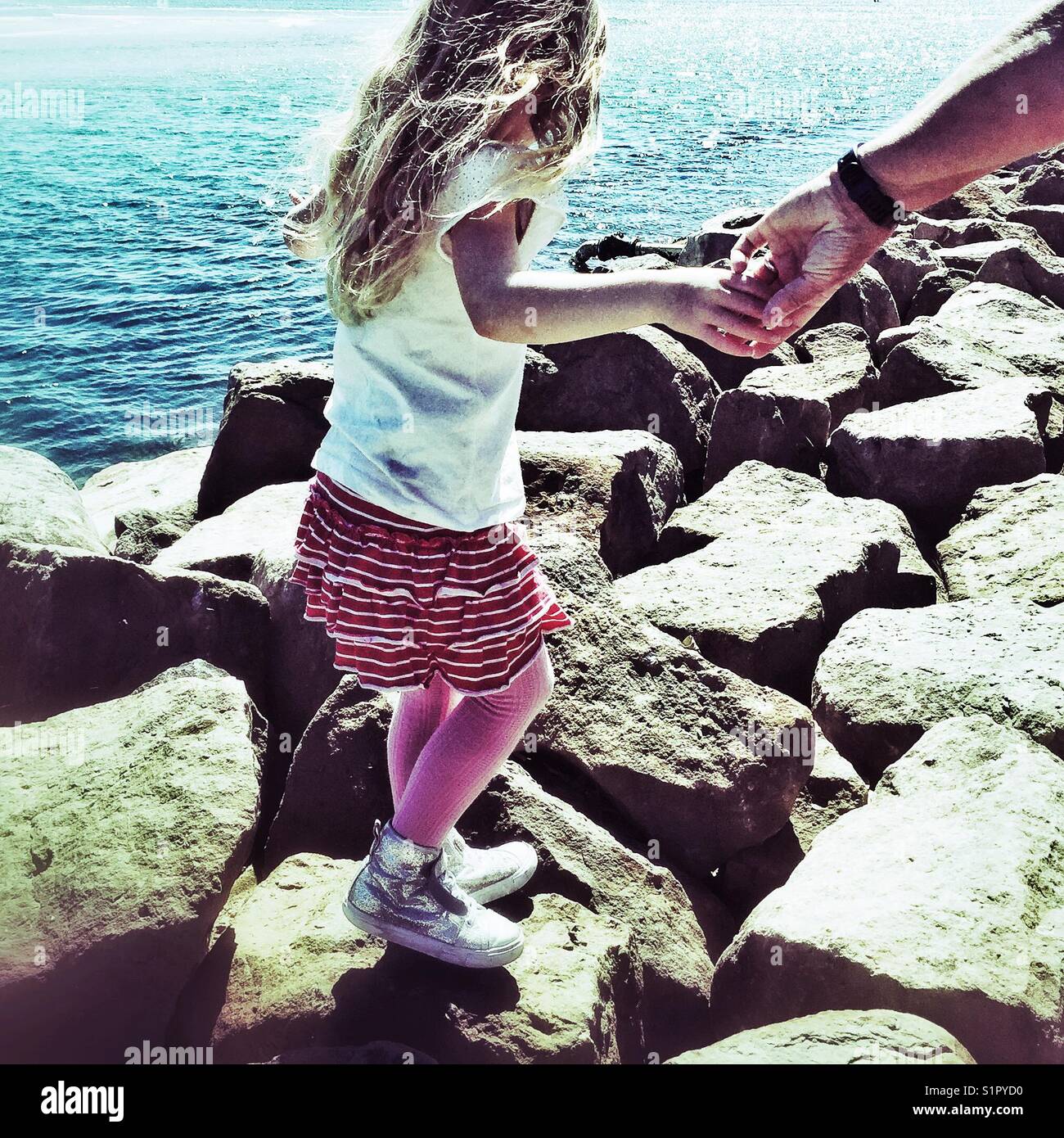 Young girl holding hands with adult walks on rocks by water edge. Huskisson, Jervis Bay, New South Wales, Australia Stock Photo