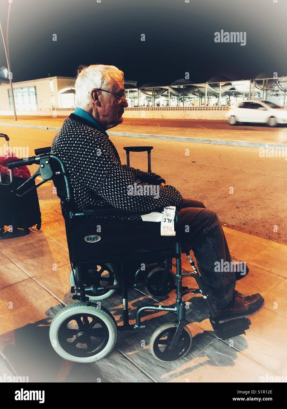 Senior man in a wheelchair waiting for a shuttle bus outside Alicante airport at night. Spain - Smartphone Captured Stock Image