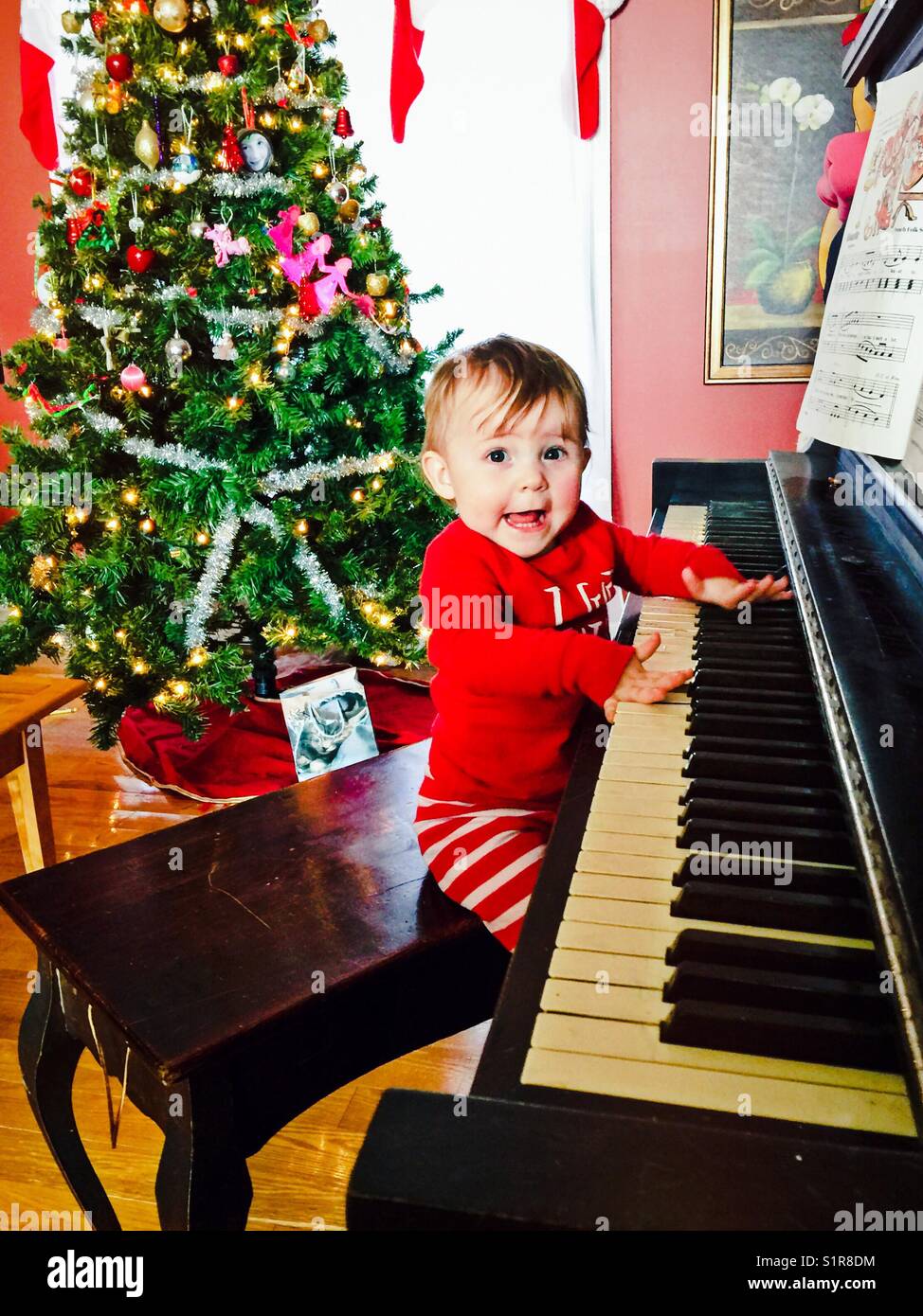 Toddler happily playing piano in Christmas pyjamas with Christmas tree in background - Smartphone Captured Stock Image