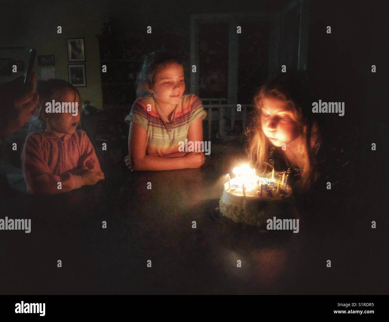 Girl blowing out the candles on her birthday cake with sisters watching Stock Photo