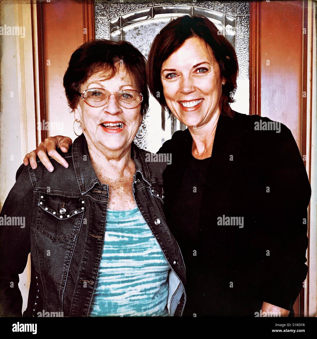 A smiling baby boomer daughter and her smiling mother face the camera in front of the exterior door. Stock Photo