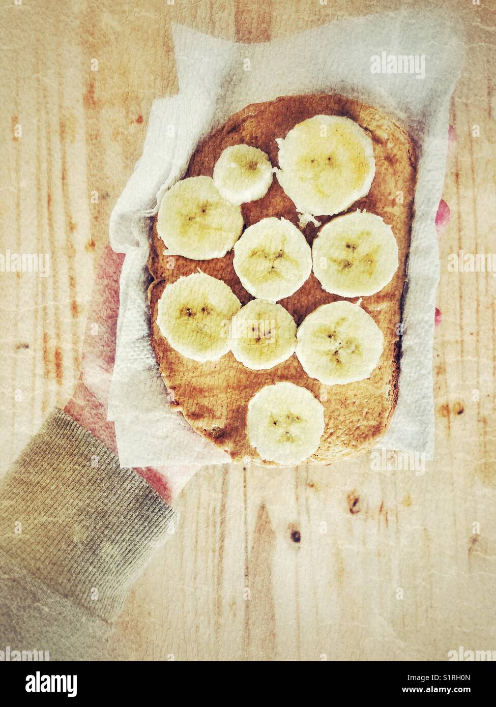 Sliced banana and peanut butter on rye toast in paper towel being held by person over wooden table Stock Photo