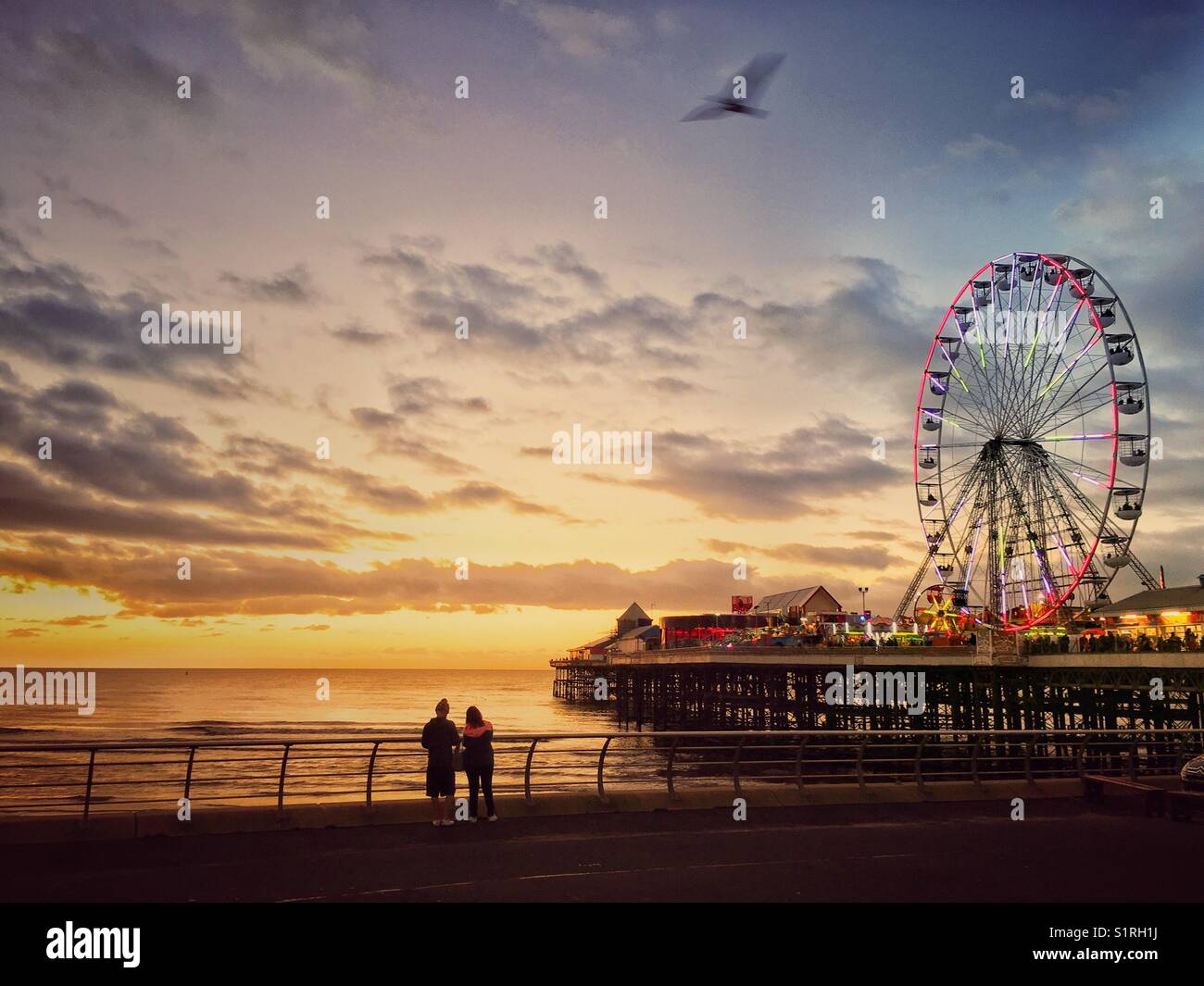 Two people looking out to sea from Blackpool promenade at sunset. Colourful sky and illuminated big wheel on Central Pier - Smartphone Captured Stock Image
