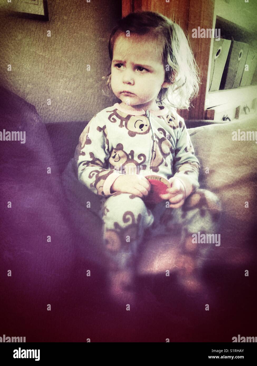 Toddler girl in monkey pyjamas sitting on a chair with a grumpy frown - Smartphone Captured Stock Image