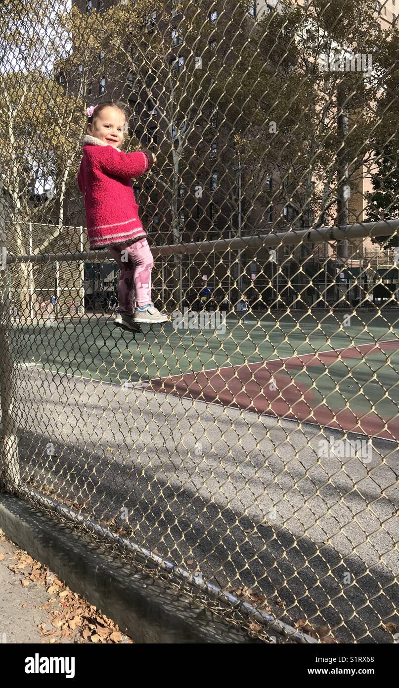Two year old girl has climbed a chicken wire fence while enjoying herself on a school playground. Stock Photo