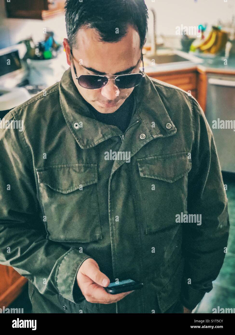 Man standing in kitchen wearing coat and sunglasses using a smartphone - Smartphone Captured Stock Image