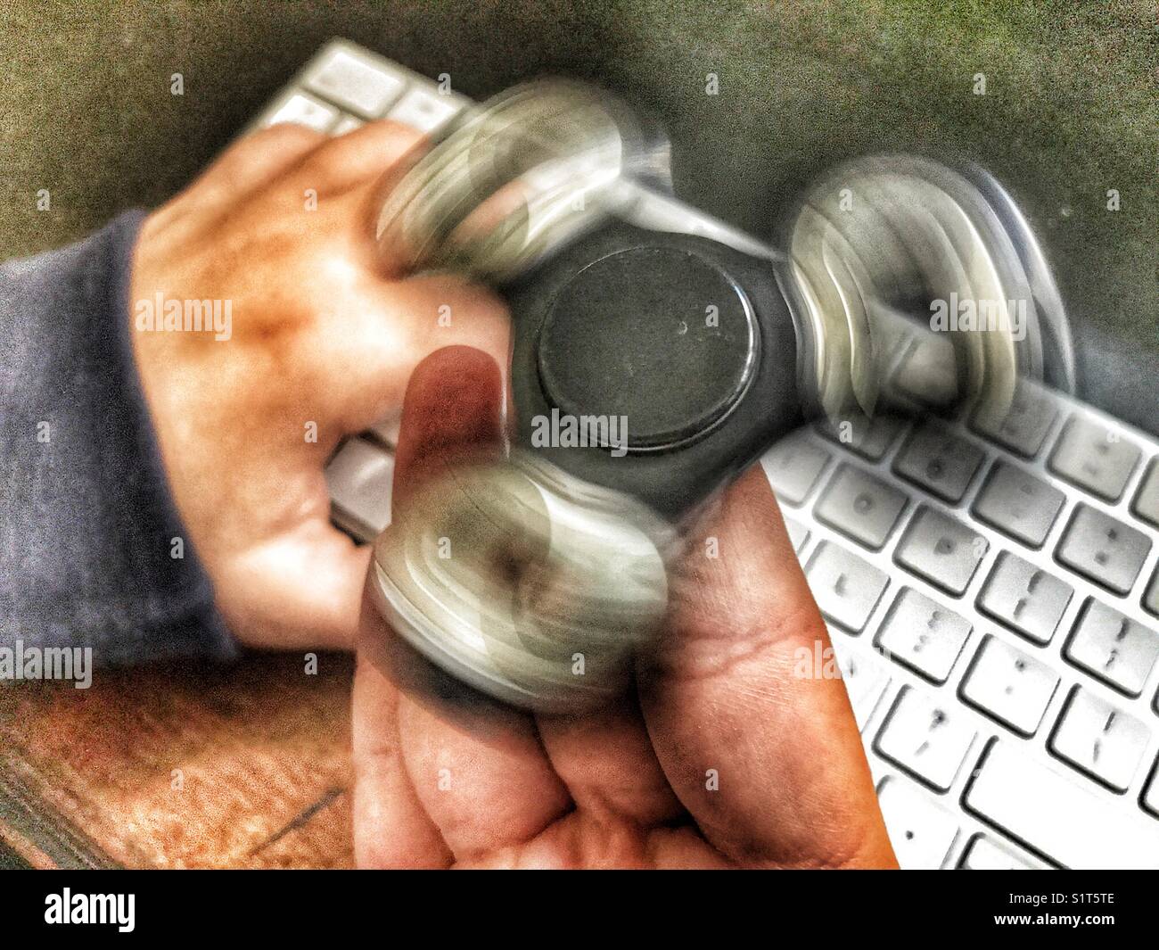 Fidget spinner, spinning on a milennial’s  hand, whilst sitting at a leather topped desk with one hand on a wireless computer keyboard. - Smartphone Captured Stock Image