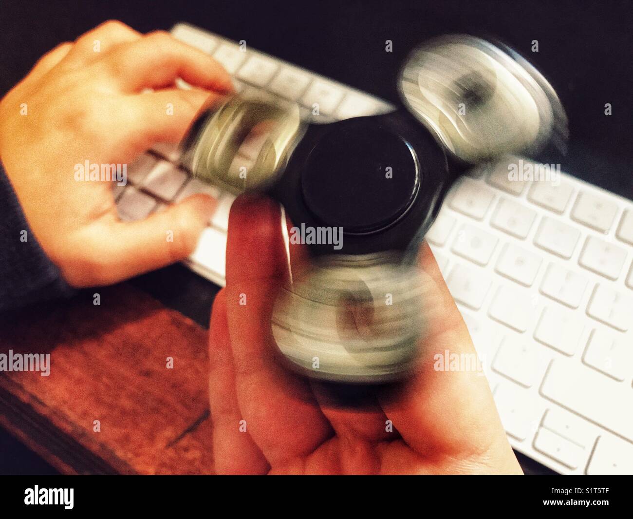 Fidget spinner, spinning on a milennial’s  hand, whilst sitting at a leather topped desk with one hand on a wireless computer keyboard. - Smartphone Captured Stock Image