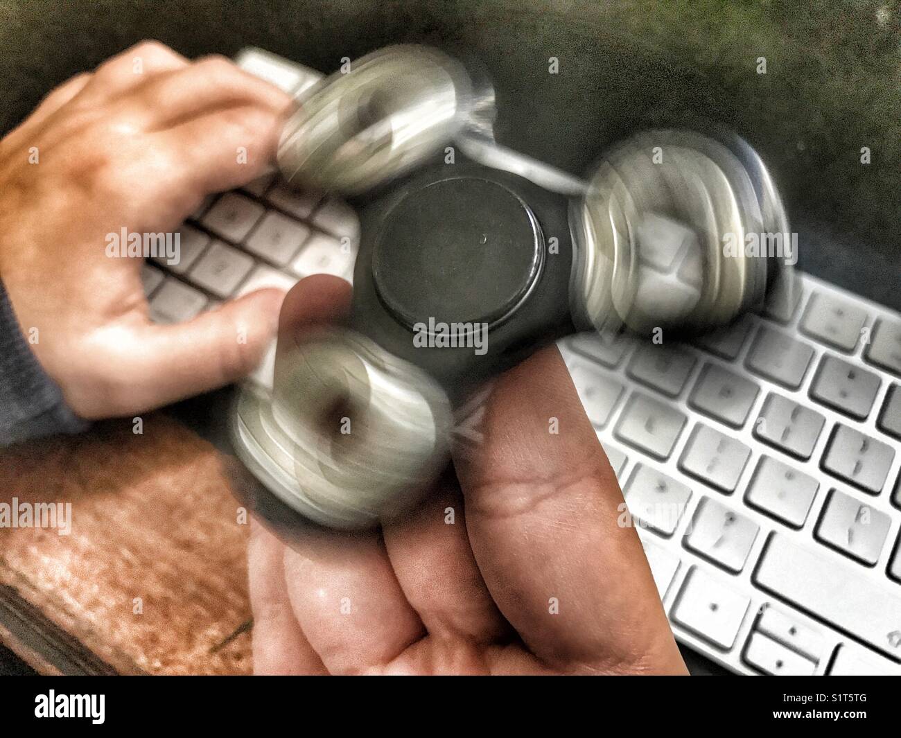 Fidget spinner, spinning on a milennial’s  hand, whilst sitting at a leather topped desk with one hand on a wireless computer keyboard. - Smartphone Captured Stock Image