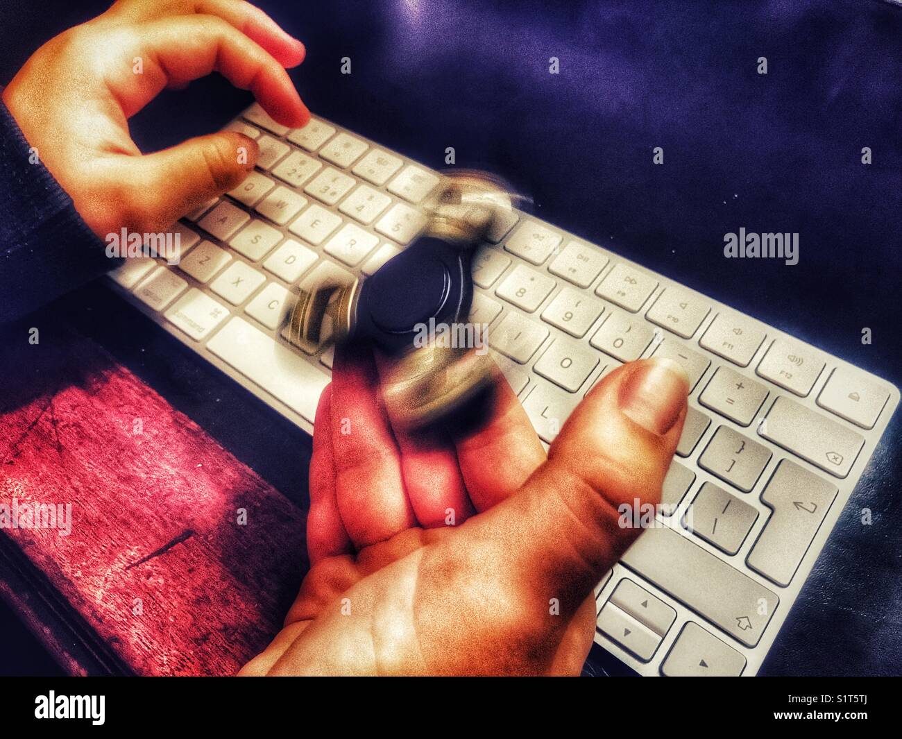 Fidget spinner, spinning on a milennial’s  hand, whilst sitting at a leather topped desk with one hand on a wireless computer keyboard. - Smartphone Captured Stock Image