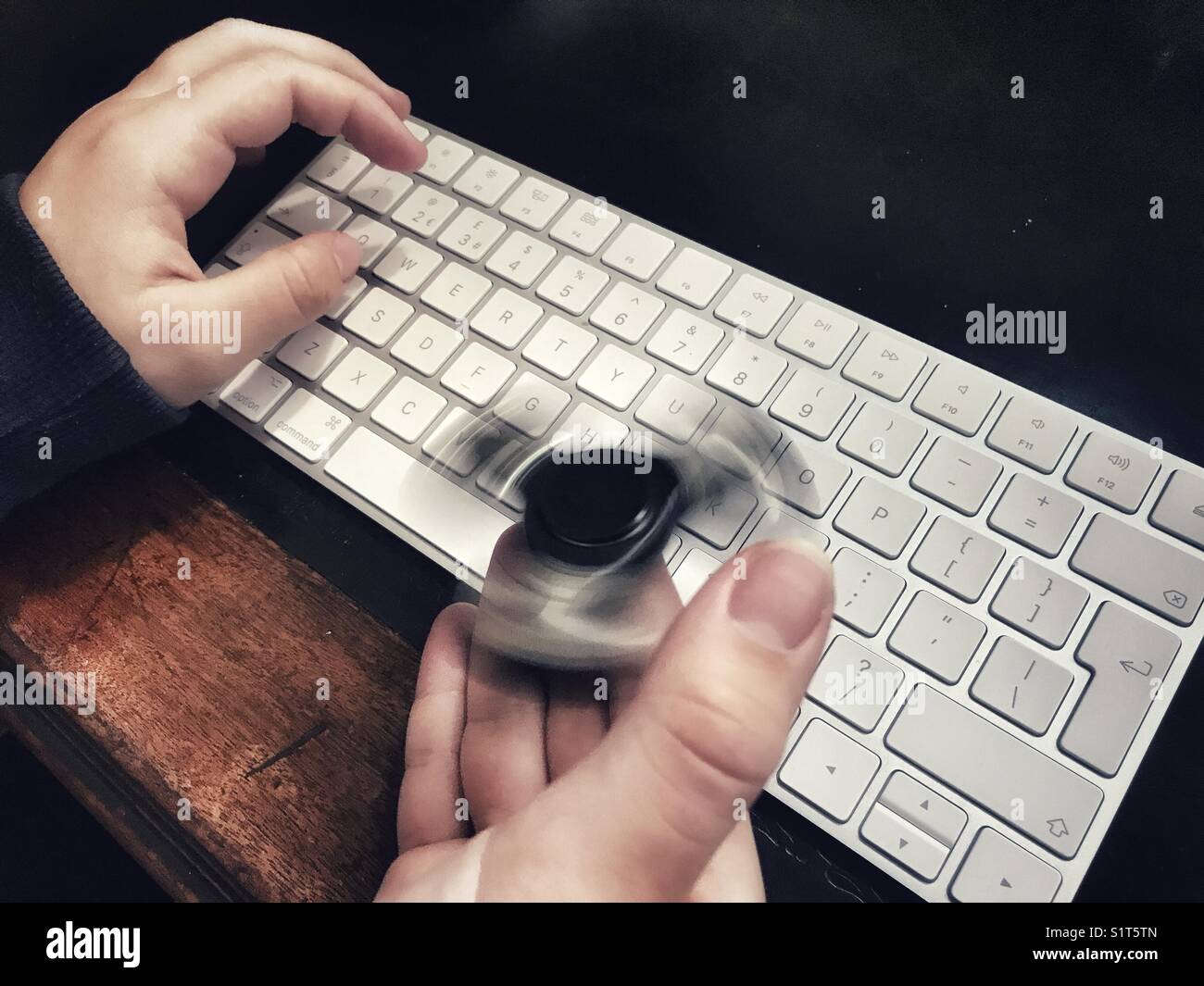 Fidget spinner, spinning on a milennial’s  hand, whilst sitting at a leather topped desk with one hand on a wireless computer keyboard. - Smartphone Captured Stock Image