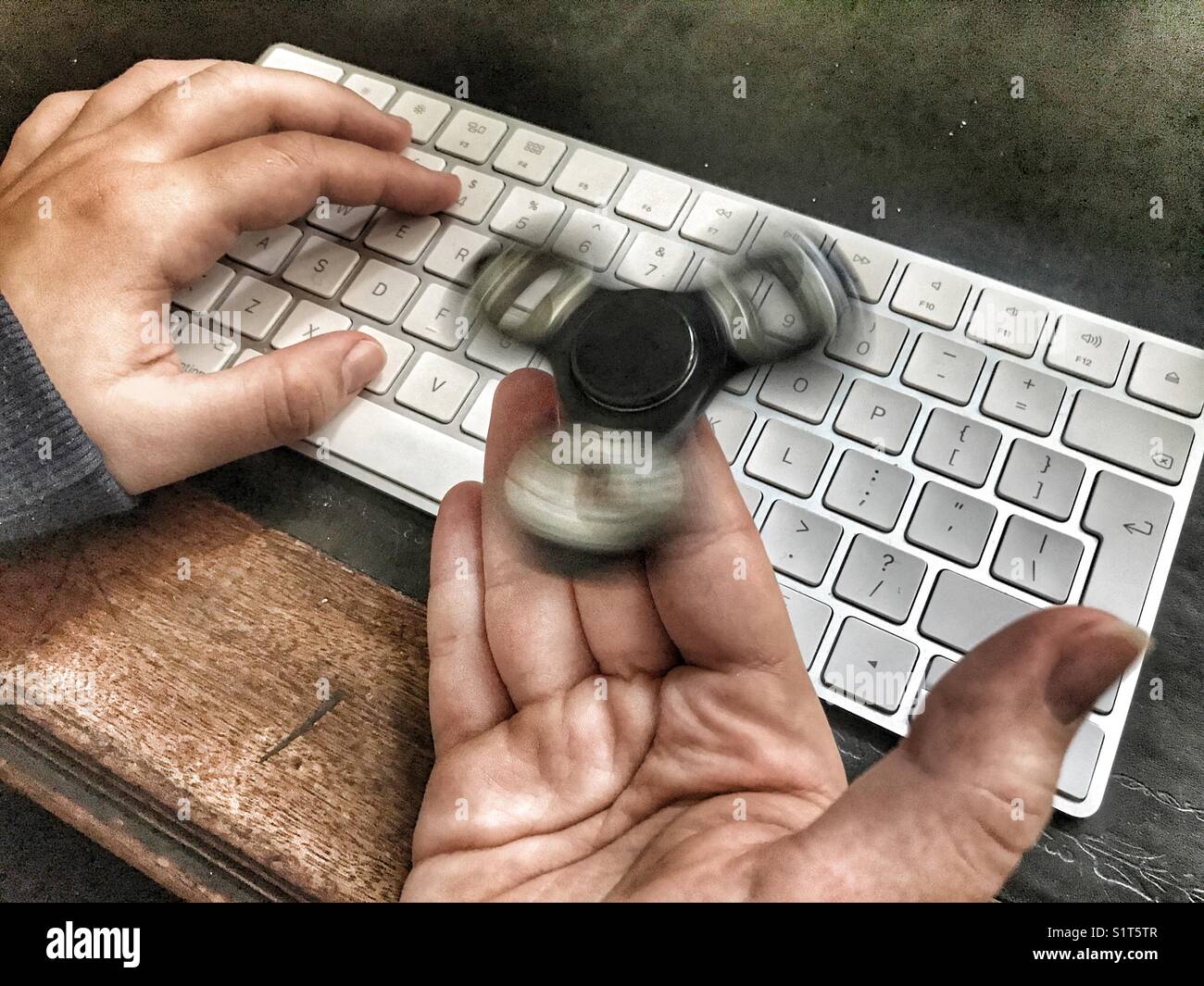 Fidget spinner, spinning on a milennial’s  hand, whilst sitting at a leather topped desk with one hand on a wireless computer keyboard. - Smartphone Captured Stock Image