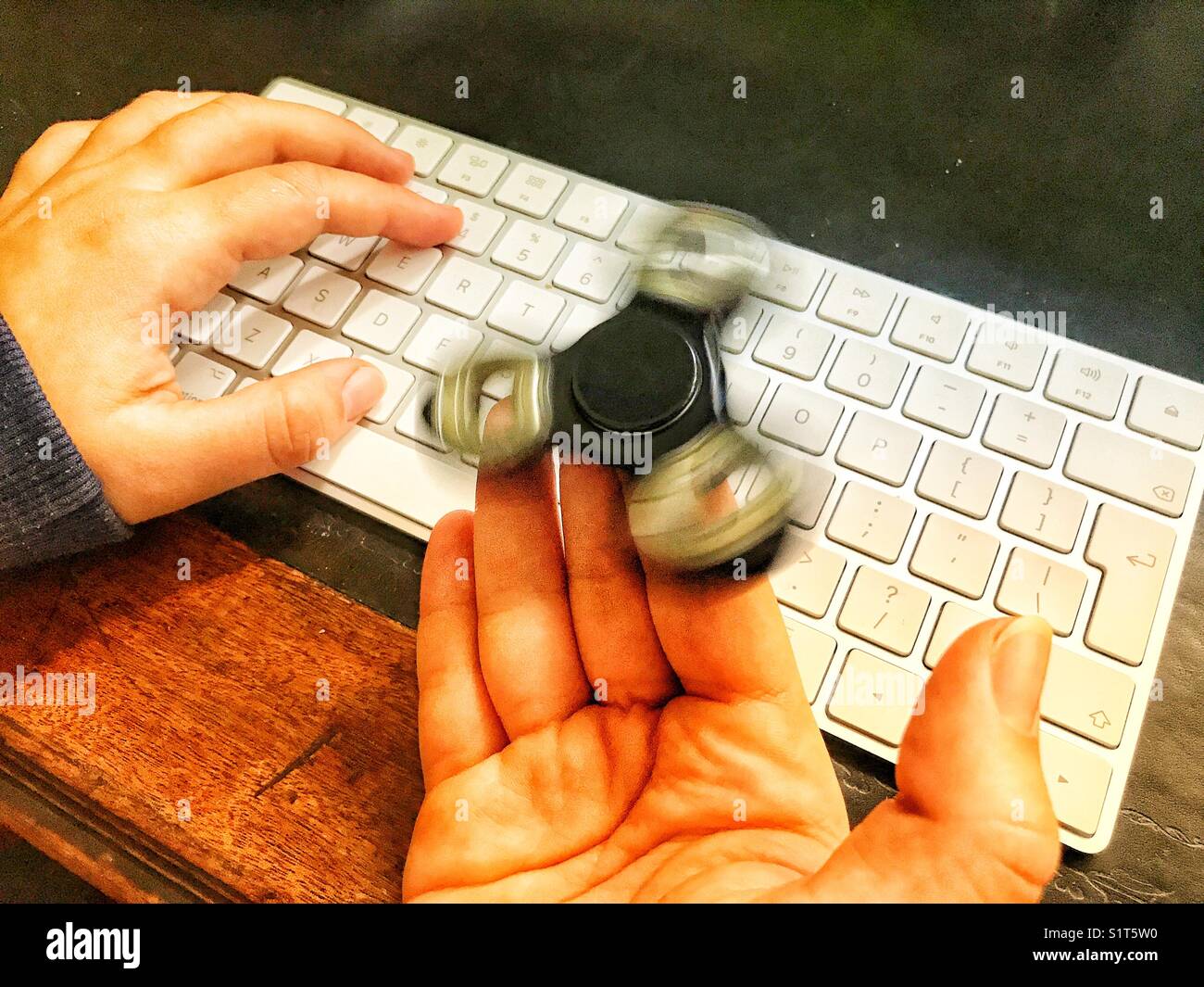 Fidget spinner, spinning on a milennial’s  hand, whilst sitting at a leather topped desk with one hand on a wireless computer keyboard. - Smartphone Captured Stock Image