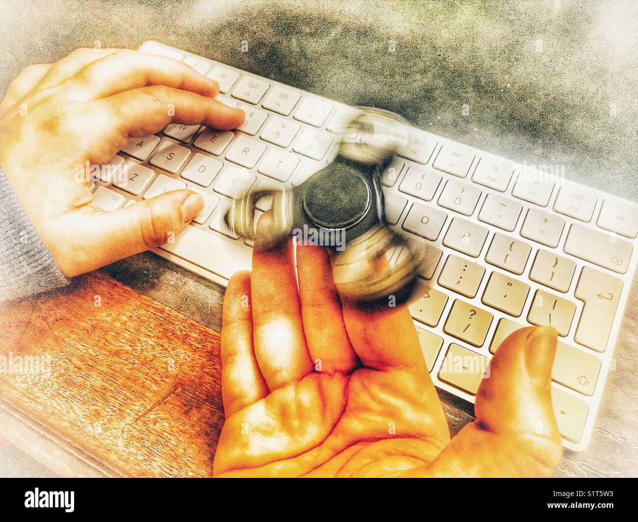 Fidget spinner, spinning on a milennial’s  hand, whilst sitting at a leather topped desk with one hand on a wireless computer keyboard. - Smartphone Captured Stock Image
