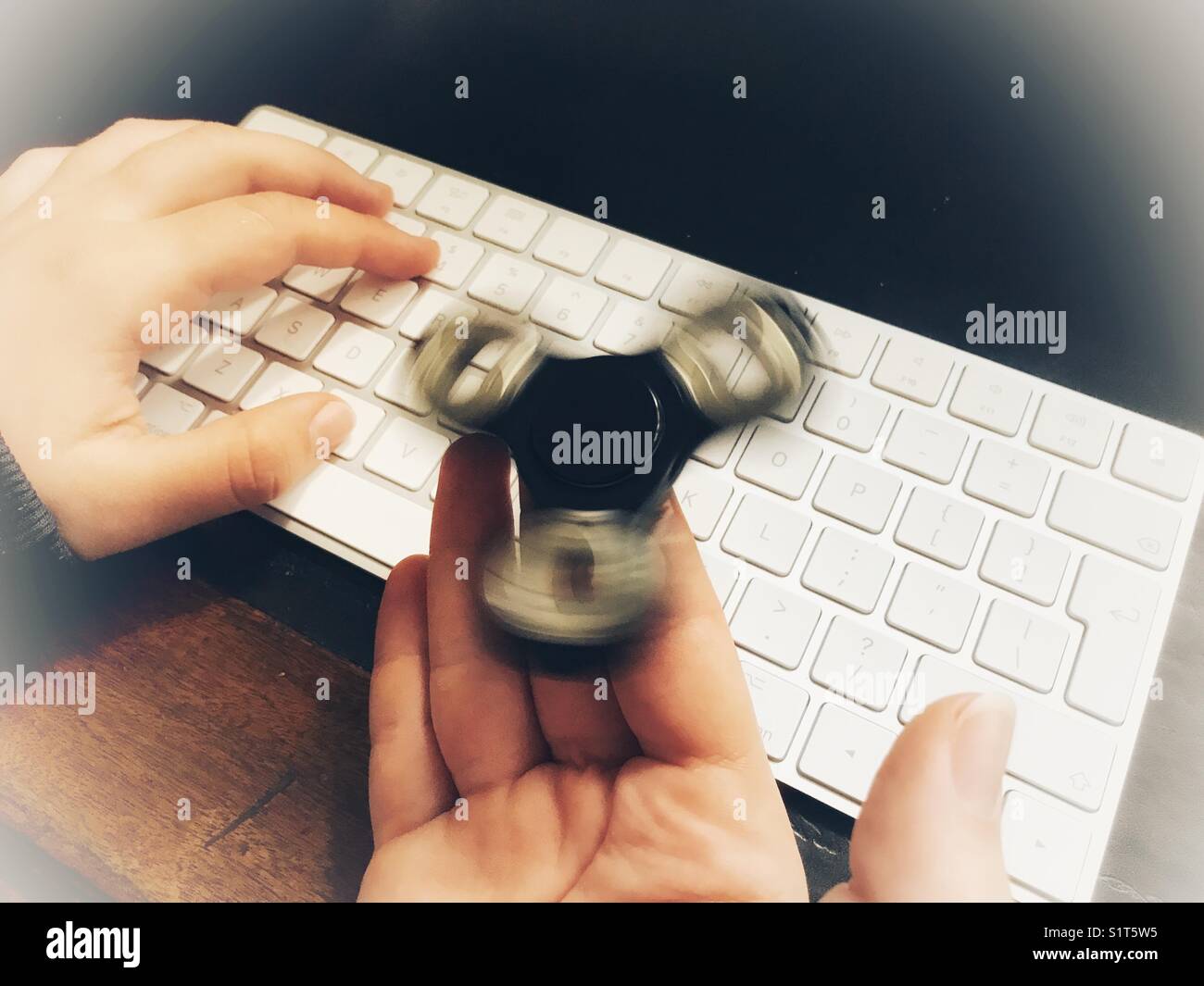 Fidget spinner, spinning on a milennial’s  hand, whilst sitting at a leather topped desk with one hand on a wireless computer keyboard. - Smartphone Captured Stock Image