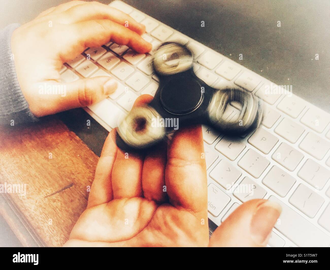Fidget spinner, spinning on a milennial’s  hand, whilst sitting at a leather topped desk with one hand on a wireless computer keyboard. - Smartphone Captured Stock Image