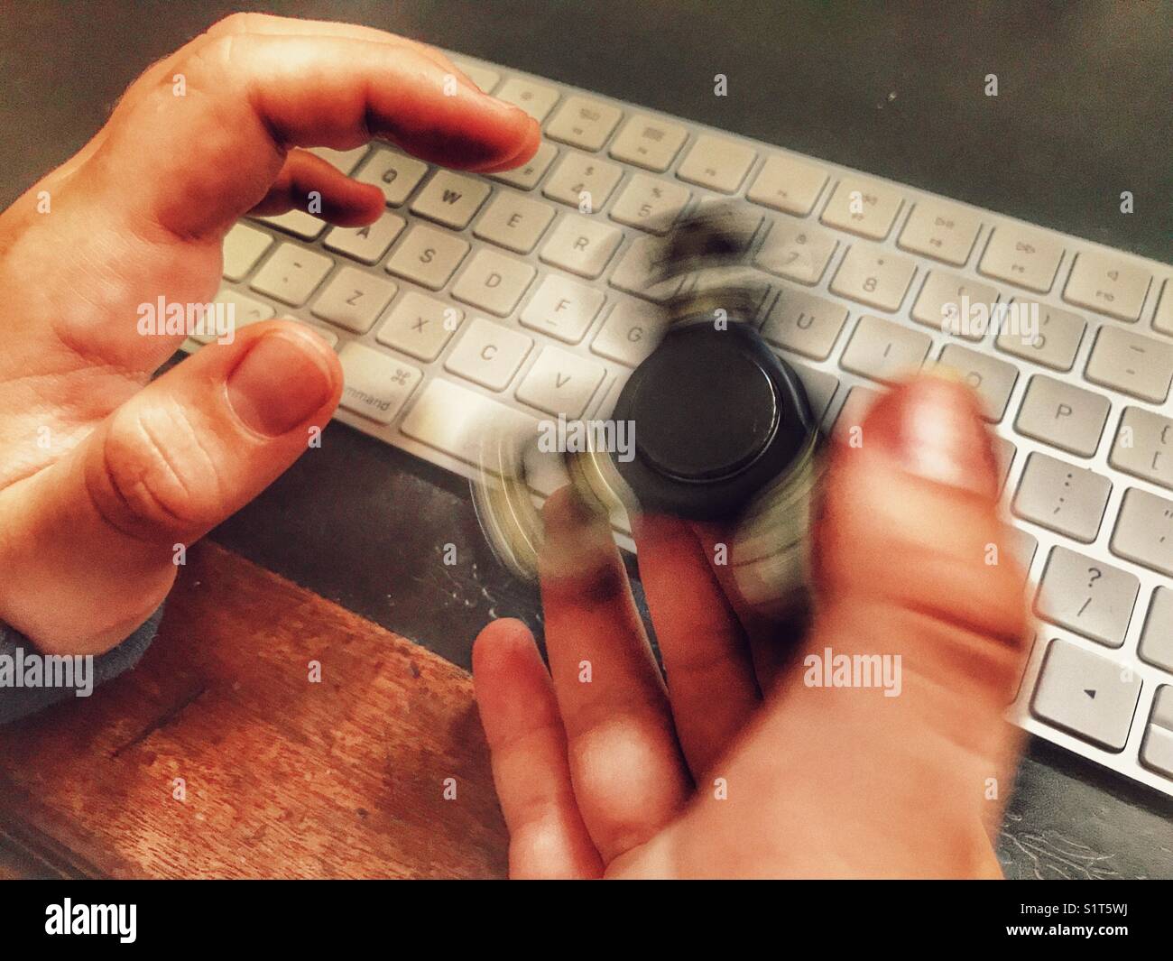 Fidget spinner, spinning on a milennial’s  hand, whilst sitting at a leather topped desk with one hand on a wireless computer keyboard. - Smartphone Captured Stock Image
