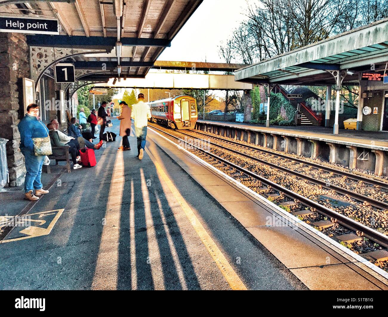 People and shadows on the rail station platform with a train approaching. Sherborne, Dorset, England - Smartphone Captured Stock Image