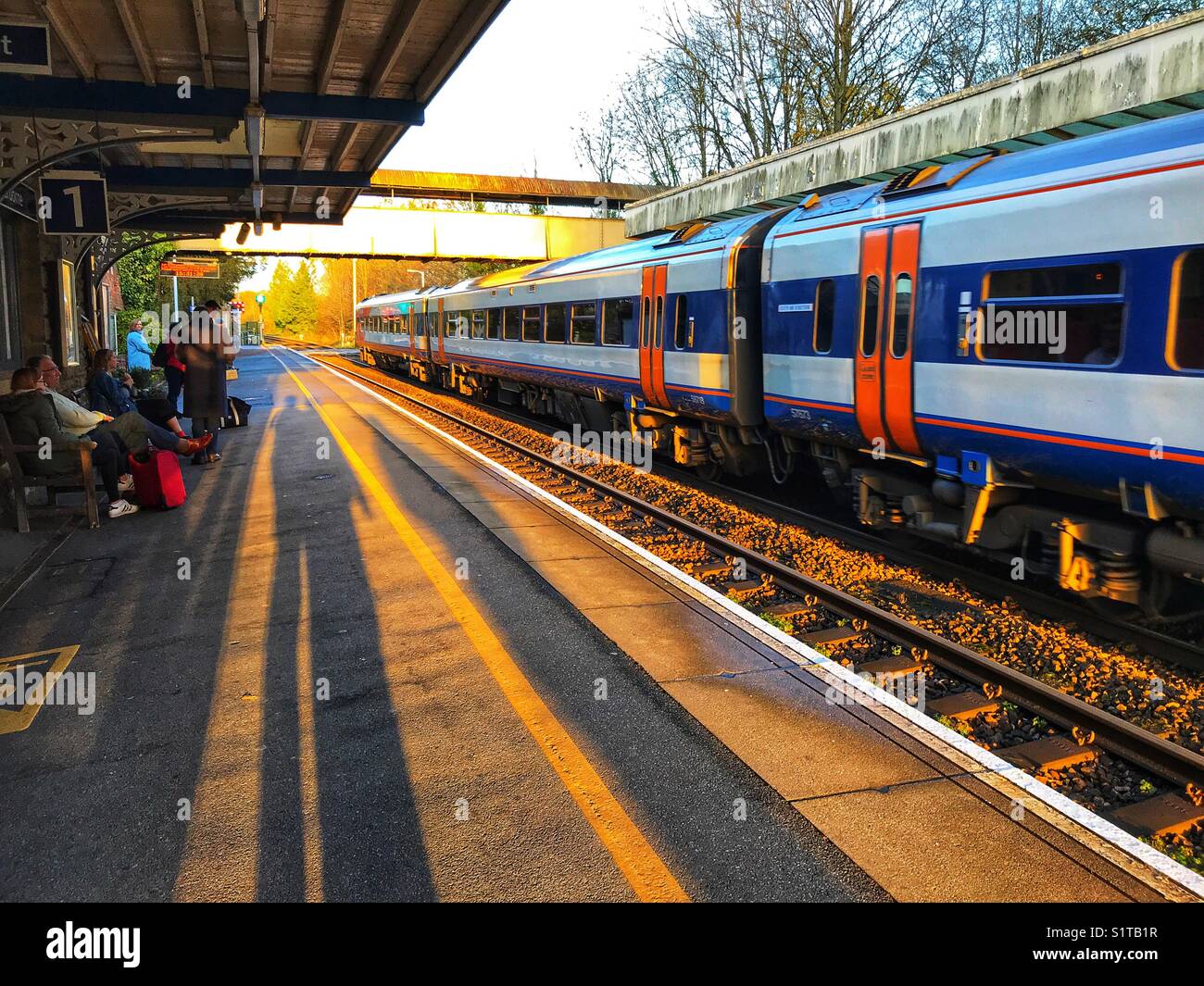 People and shadows on the rail station platform with a train on the opposite platform. Sherborne, Dorset, England - Smartphone Captured Stock Image