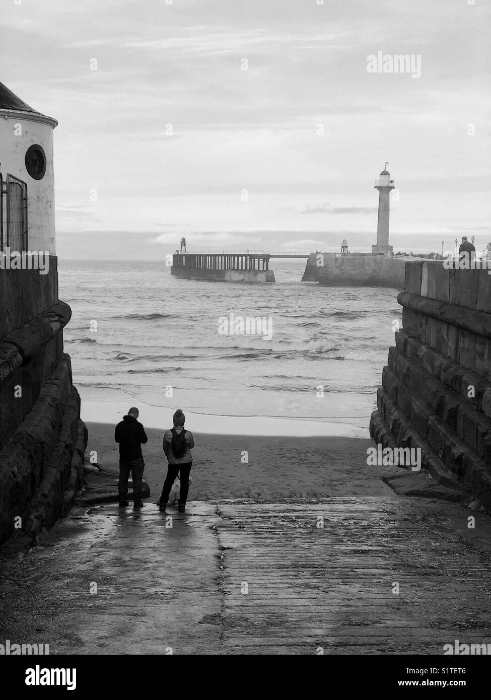 Couple standing on a slipway watching the waves rolling in at Whitby, UK - Smartphone Captured Stock Image