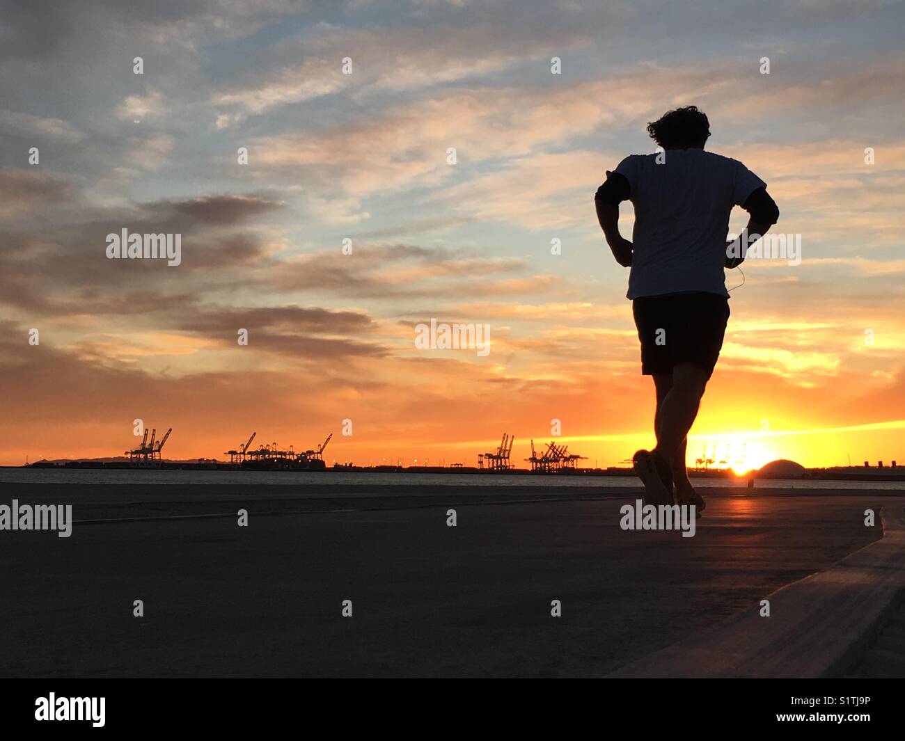 Man running on the beach at sunset with dramatic clouds and the Port of Long Beach, CA, USA in the background. - Smartphone Captured Stock Image