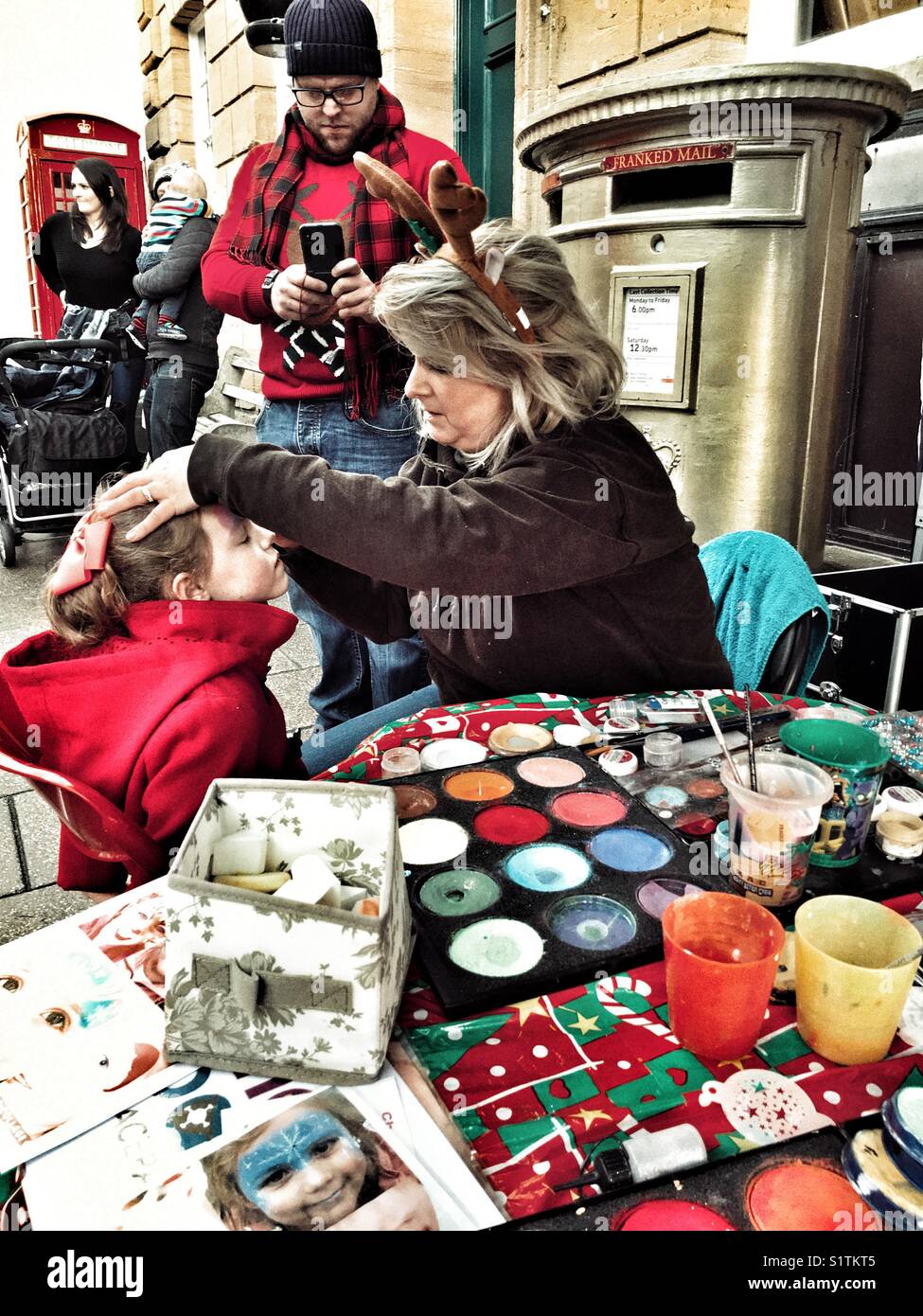 Man taking photo on his phone of child having her face painted, outdoors - Smartphone Captured Stock Image