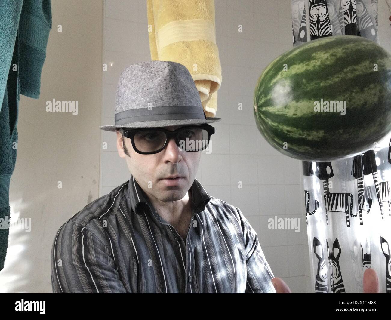 Portrait of man with dark glasses and hat in toilet with watermelon in the air - Smartphone Captured Stock Image