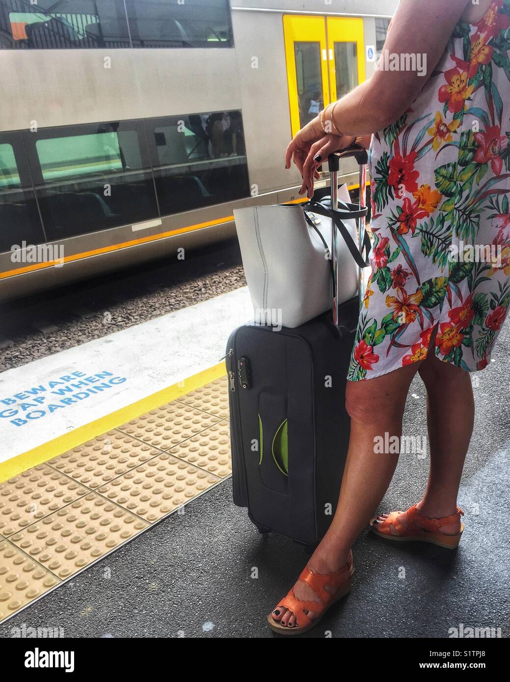 Stylish woman stands waiting on train station platform with wheeled suitcase. Travellers summer vacation train journey. Lady in bright dress waits at railway station with luggage. Stock Photo