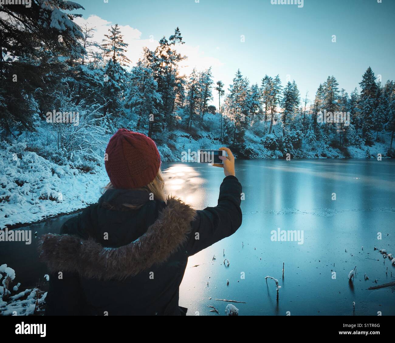 Woman wearing a dark red knitted hat and thick winter jacket taking a photograph of a frozen lake with a smart phone. - Smartphone Captured Stock Image