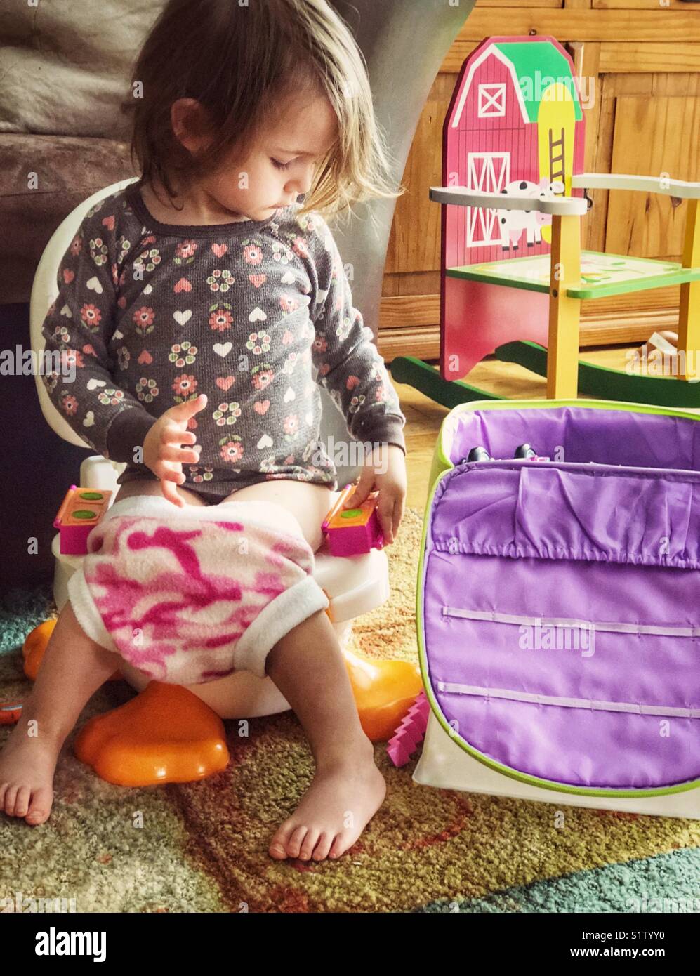 Toddler Girl Sitting On Potty Chair In Family Room Playing With Toys 