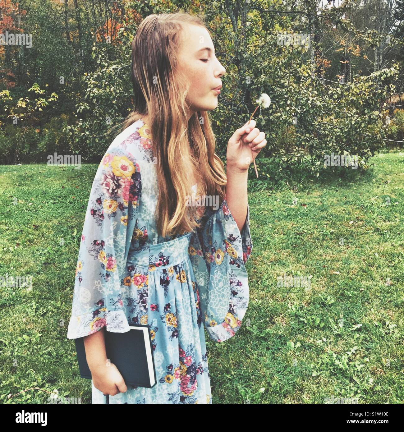Girl wearing vintage 60’s era dress blowing on fluffy white dandelion outside - Smartphone Captured Stock Image