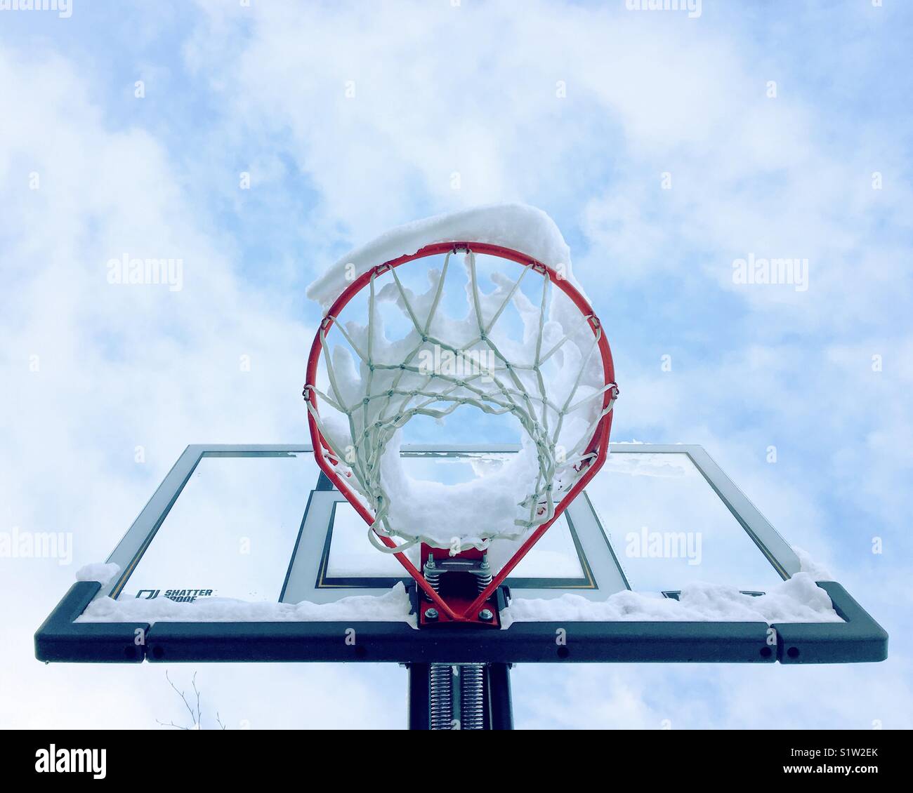 Looking up at a snow covered basketball hoop with little white clouds and blue sky above. Stock Photo