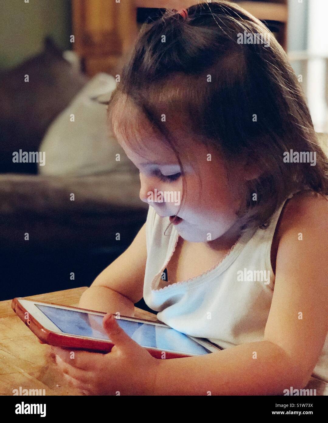 Two year old toddler girl wearing white undershirt playing with tablet in pink case at a wooden table - Smartphone Captured Stock Image