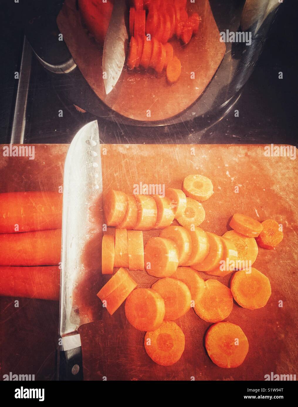 From above view of chopping fresh carrots with large chef’s knife on a brown cutting board with reflection in stainless steel pot above Stock Photo