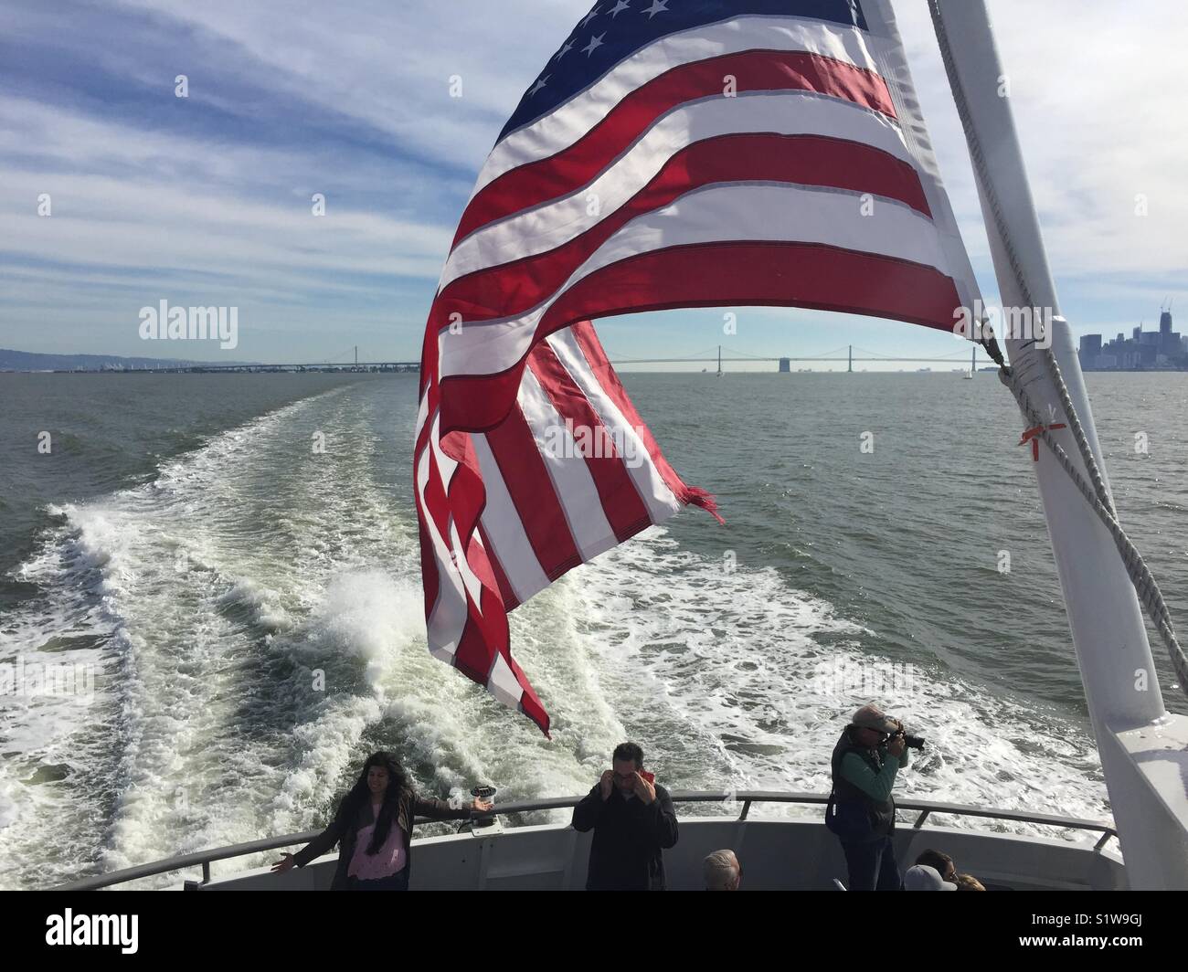 Ferry going from San Francisco to Sausalito.  American flag in foreground.  Bay Bridge and San Francisco skyline in background. Stock Photo