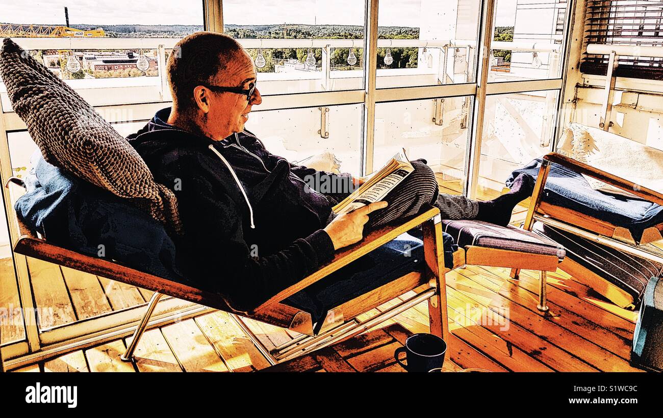Middle aged Caucasian man lying on reclining chair on enclosed terrace reading a book, Sweden Stock Photo